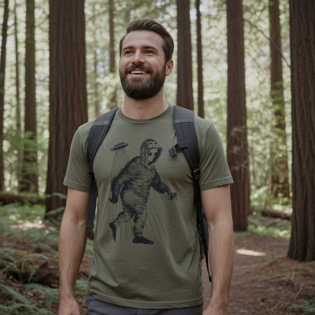Man wearing a olive green t-shirt with alien bigfoot in a forest